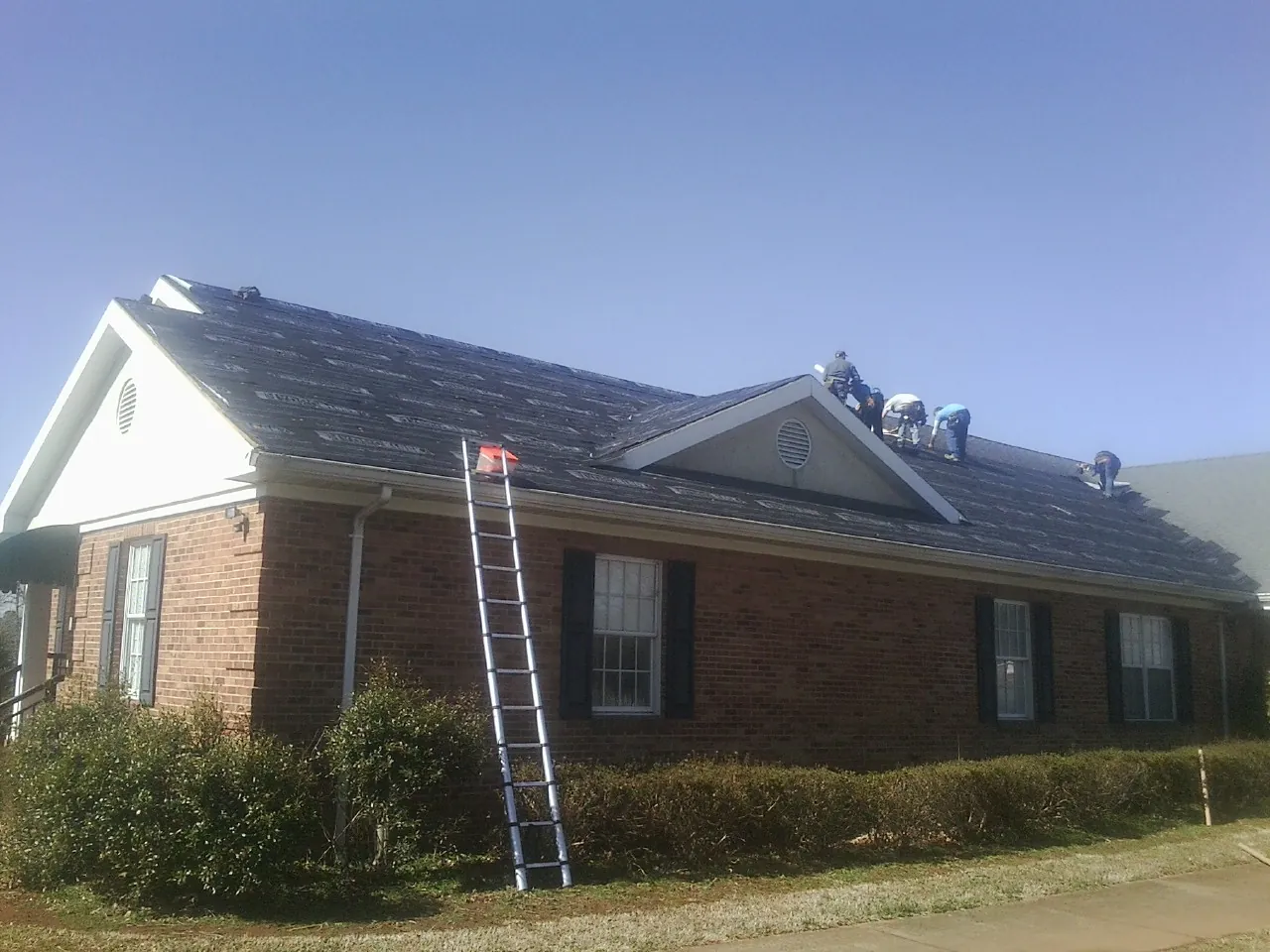Crew tearing shingles off a brick ranch roof with a ladder set — Metal Roof Repair work by our crew in Ellis