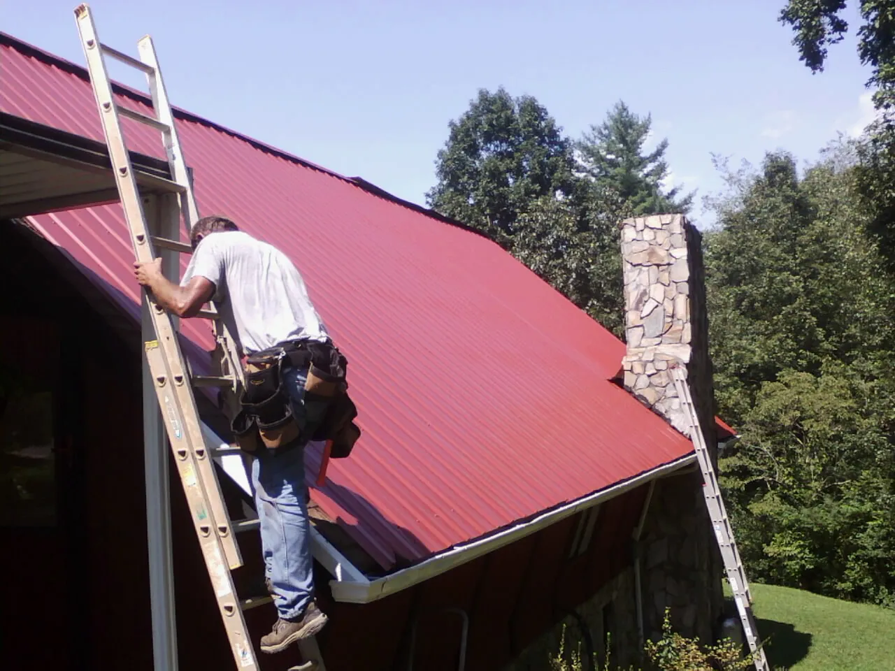 Roofer on a ladder beside a red metal roof and stone chimney — Soffit & Fascia Repair by Roofing Brothers in Ellis